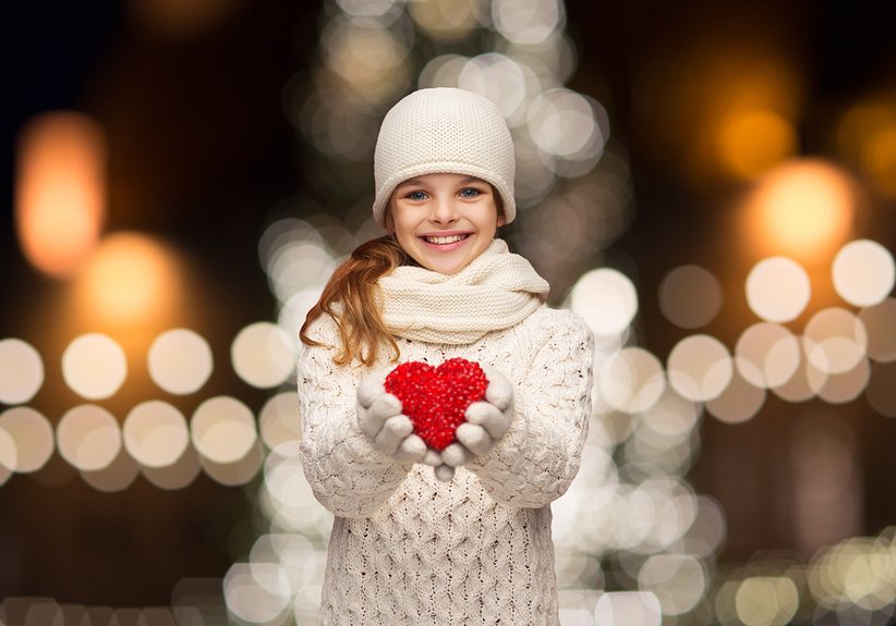 love, charity, holidays and people concept - happy girl in winter clothes with red heart over christmas tree lights background